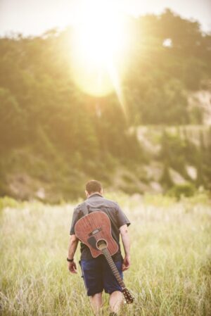 Vertical shot of a male walking in a field next to a forest with a guitar on his back