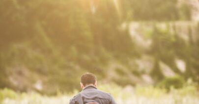 Vertical shot of a male walking in a field next to a forest with a guitar on his back